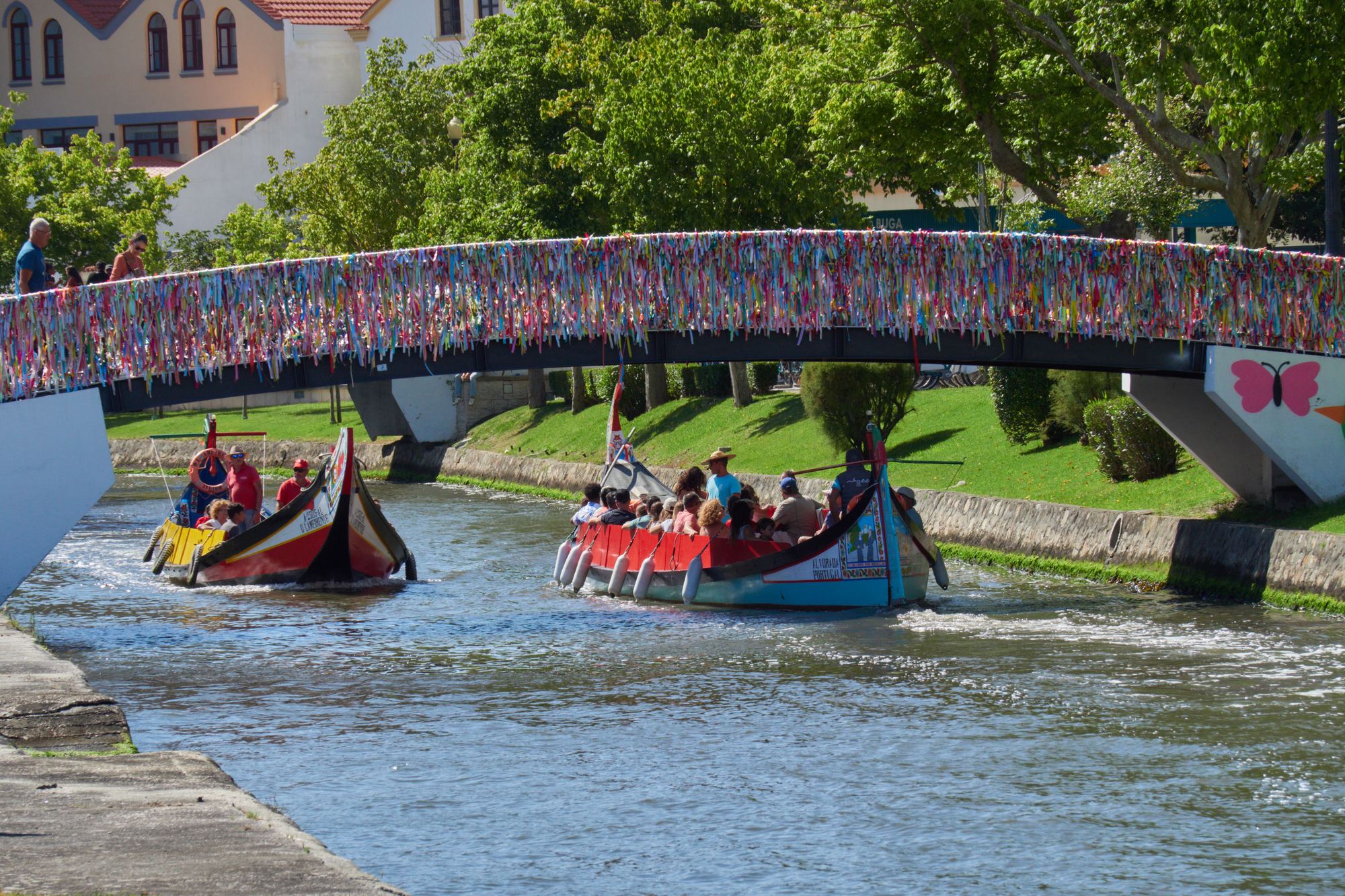 Dois barcos moliceiros com passageiros debaixo de uma ponte pedonal cheia de fitas, num canal de Aveiro