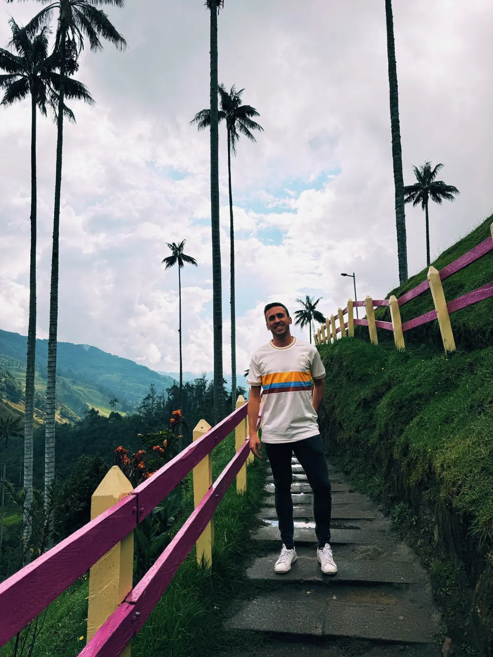 Alberto sonríe de pie en un sendero de piedra del Valle de Cocora, con una baranda rosada y amarilla a un lado y palmas de cera altísimas contra un cielo nublado