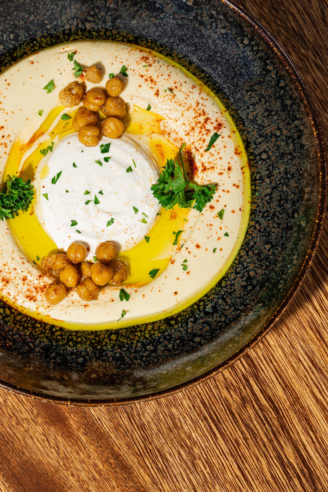 Hummus con crema blanca al centro, garbanzos crocantes, aceite y perejil en plato oscuro